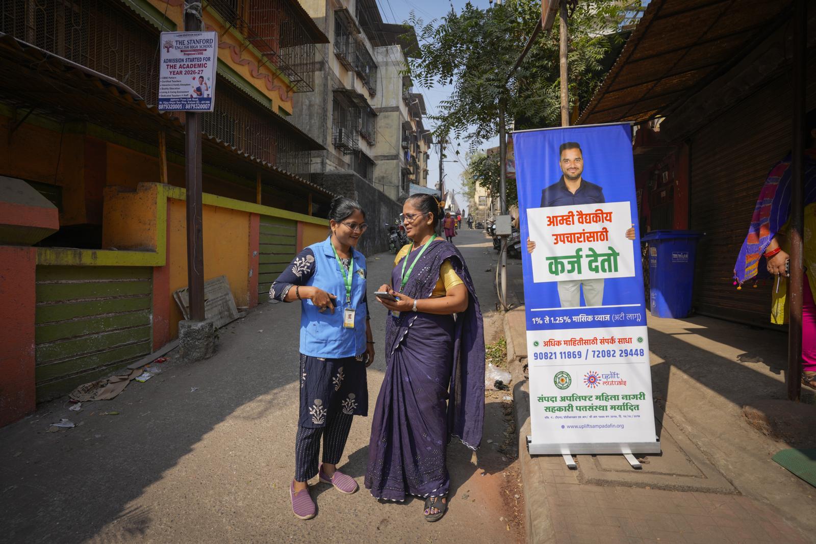 Two health financing officers standing in a street next to a Sampada banner