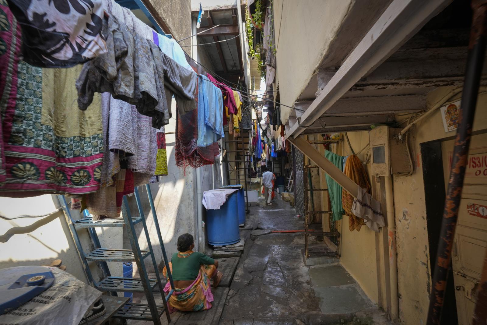 A narrow lane between houses in a Mumbai settlement, with washing hung overhead and a woman seated outside her door